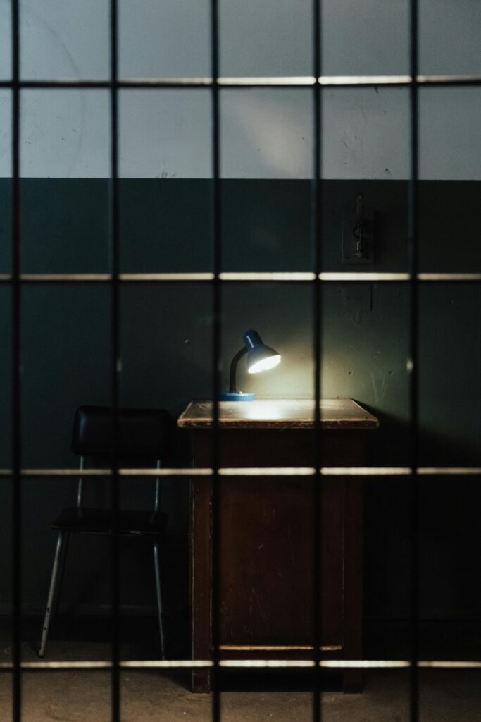 A dimly lit prison cell with a lamp on a table behind bars.